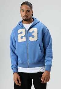 Blauwe zip-up hoodie van zacht stof, met een grote witte verweerde "23" op de voorkant. Voorzien van een voorzak en capuchon.