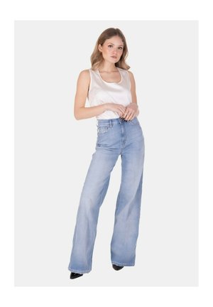 Giovane donna in piedi che indossa un top in raso bianco senza maniche e jeans azzurri a vita alta con gambe larghe e scarpe nere con punta.