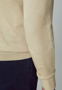 Beige trui met geribbelde manchetten, voorzien van genaaide elleboogpatches in een honingraatpatroon. Gladde stof met een getailleerde snit.