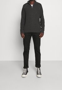 Mann in einem dunkelgrauen Strickpullover mit halben Reißverschluss, schwarzen umgeschlagenen Hosen und schwarzen hoch geschnittenen Sneakers mit weißen Sohlen und Schnürsenkeln, stehend.