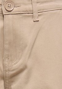 Pantalon en coton beige avec une texture lisse, présentant une fermeture à bouton, des détails de couture et des poches latérales.