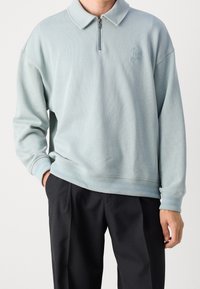 Ljusblå pullovertröja med dragkedja i kragen, ribbstickade muddar och nederkant, med en diskret logotyp på bröstet. Tillverkad av mjukt material.