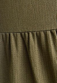 Tissu texturé vert olive présentant un motif de froissage subtil avec des détails froncés et ruchés le long de la couture.