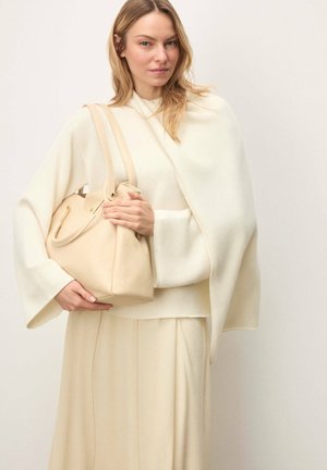 Femme aux cheveux blonds portant une tenue crème tient un sac à main en cuir beige sur un fond uni clair.