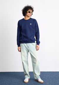 Mørkeblå sweatshirt med rund hals og lille hvidt logo, parret med lyseblå løstsiddende jeans og hvide sneakers med røde accenter.