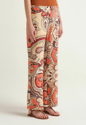 Wide-leg pants with intricate orange, beige, and brown paisley and floral patterns, worn with brown thong sandals and an orange top.