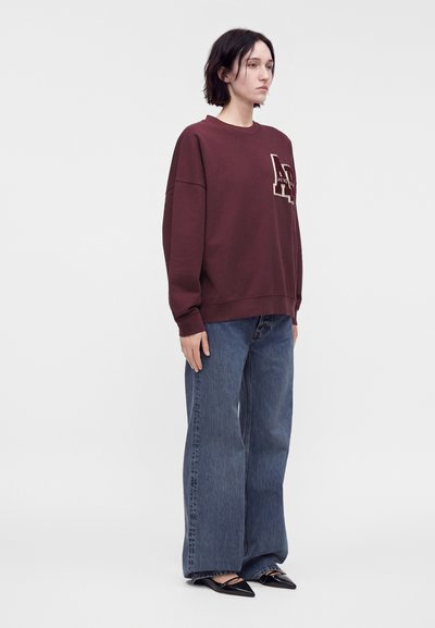 Γυναίκα που στέκεται, φορώντας ένα μπορντό oversized πουλόβερ με γράμμα σχεδίου, φαρδιά μπλε τζιν και μαύρα μυτερά επίπεδα παπούτσια.