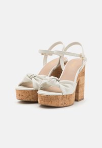 Sandales à plateforme blanches avec des talons en liège, présentant un design à l'avant noué et une bride de cheville avec une boucle dorée. Texture lisse sur le dessus.