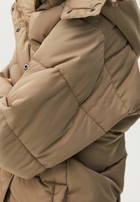 Doudoune marron avec de grandes sections matelassées, une fermeture éclair sur le devant et des boutons-pression sur le col. Texture douce avec une finition légèrement brillante.