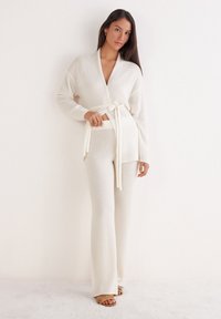 Femme portant un pull en tricot crème noué à la taille et un pantalon large assorti, se tenant devant un mur blanc avec des sandales.