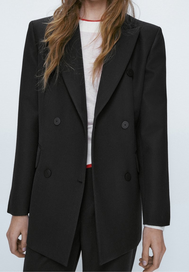 Femme portant un blazer noir croisé sur un haut blanc avec une bordure rouge, debout devant un fond clair uni.