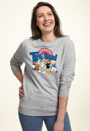 Femme souriante portant un sweat-shirt gris avec des personnages de dessin animé colorés de Tune Squad et un jean bleu, debout devant un fond uni.