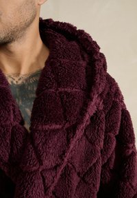 Nærbillede af en person iført en textured dyb bordeaux fleecebadekåbe med sjalskrave, der afslører en brysttatovering af en ugle.