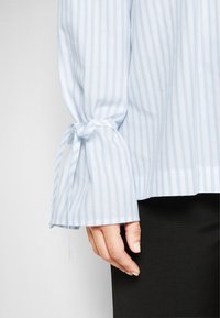 Světle modrá pruhovaná blůza s dlouhými rukávy a detailem vázání na manžetách. Tkanina vypadá hladce a lehce, kombinována s tmavými kalhotami.