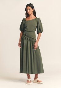 Abito maxi verde oliva con maniche a palloncino, scollo quadrato, corpino arricciato e gonna fluida. Indossato con sandali platform color crema.