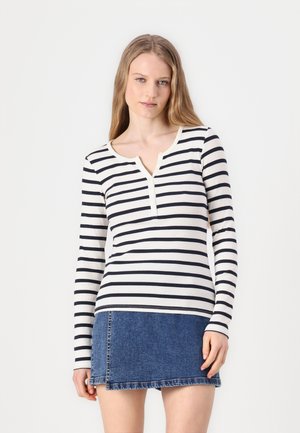 Haut à manches longues de couleur crème avec des rayures marine, présentant un col henley et trois boutons. Associé à une jupe en jean. Tissu lisse.