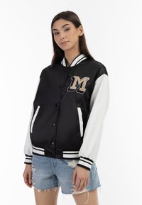 Jachetă bomber din saten negru cu mâneci albe, cu manșete ribosite și un logo „M” bej pe piept. Dispune de buzunare laterale și închidere cu capse.