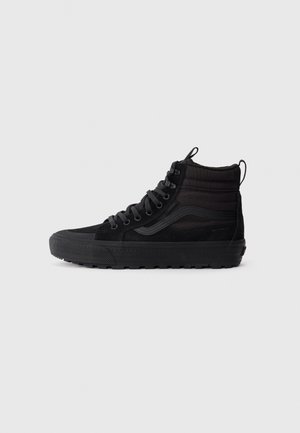 Zwarte hoge sneaker met dikke zool, veters aan de voorkant, gevoerde enkellip en waterdichte stof tegen een effen lichte achtergrond.