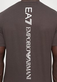 Tamsiai rudos trumpomis rankovėmis marškinėlius, kuriuose ant nugaros spausdintas vertikaliai esantis baltas Emporio Armani logotipas ir tekstas, dėvi žmogus.