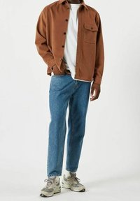 Chemise marron à boutons sur un t-shirt blanc, associée à un jean bleu et des baskets grises. Comprend une poche poitrine et une coupe décontractée.