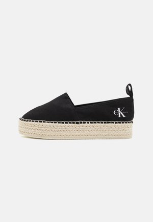 Alpargata negra sin cordones con suela gruesa de yute trenzado y logotipo de Calvin Klein Jeans en la lengüeta del talón, vista lateral sobre fondo blanco.