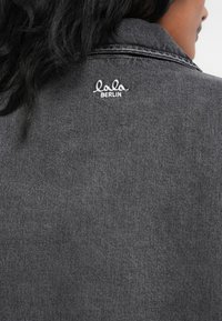 Siva denim jakna z ovratnikom, vidnimi šivi in vezenim logotipom "lala BERLIN" na hrbtu. Gladka tekstura in prilegajoča zasnova.