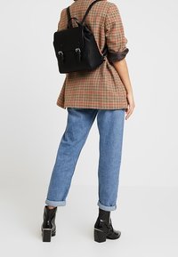 Mochila negra con herrajes plateados y correas ajustables, llevada sobre un blazer de cuadros, combinada con unos jeans azul con dobladillo enrollado y botas negras hasta el tobillo.