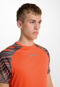 Kortärmad träningsskjorta i klar orange med ett mönstrat grått och svart axeldesign. Har logotyp och slätt, fuktavvisande tyg.