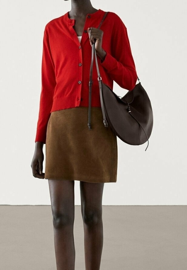 Femme portant un cardigan rouge boutonné, une mini-jupe en daim marron et un grand sac à bandoulière marron foncé, debout à l'intérieur.