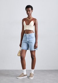 Háčkovaný bralette v krémovej farbe s červenými akcentmi, spárovaný s svetlo modrými džínsovými šortkami. Doplnky zahŕňajú malú zlatú kabelku a béžové tenisky.