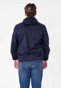Giacca impermeabile blu navy con cappuccio, caratterizzata da una texture liscia e polsini elasticizzati. Indossata con jeans blu, vista da dietro.