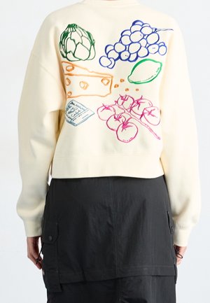 Sweat-shirt couleur crème avec des légumes brodés colorés, du fromage, des raisins, un citron et un petit paquet dans le dos, porté avec un pantalon noir.