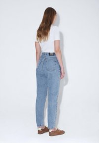 Světle modré džíny s vysokým pasem a volným střihem, dvě zadní kapsy a černá štítek na pásku, v kombinaci s bílým tričkem a hnědými botami.