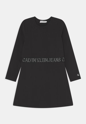 Zwarte jurk met lange mouwen en ronde hals, met een "Calvin Klein Jeans" logoband rond de taille en een klein "cK" logo op de linkermouwboord.