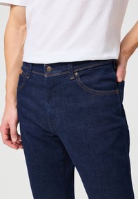 Jeans de mezclilla azul oscuro con un corte ajustado, que presentan costuras en contraste, cinco bolsillos y un cierre de botón de metal en la cinturilla.