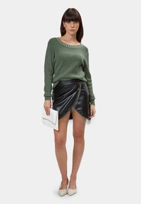 Groene gebreide trui met een decoratieve halslijn, gecombineerd met een zwarte faux leren wikkelrok met franjes en een witte clutch.
