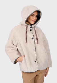 Jasno beżowa kurtka z kapturem, wykonana z miękkiego, puszystego materiału, z czarnymi guzikami i sznurkami w panterkę, o luźnym kroju i z kieszeniami.