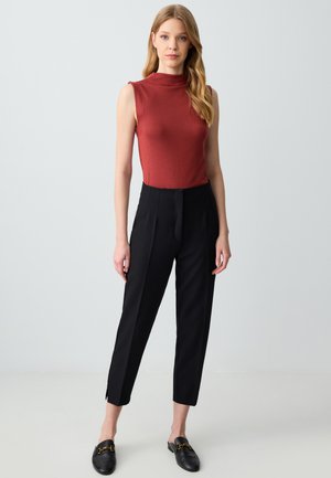 Top arrugginito senza maniche con collo alto, abbinato a pantaloni neri su misura con vestibilità slim e spacchi laterali. Mocassini neri slip-on.