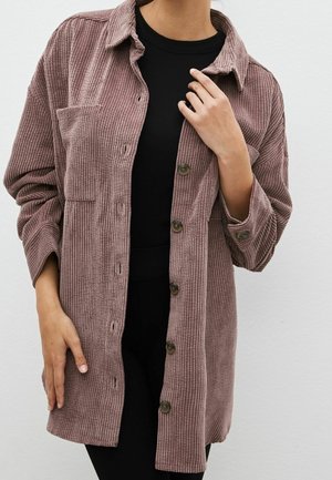 Personne portant une veste mauve en velours côtelé à boutons sur un haut noir, une main ajustant le col de la veste, l’autre reposant sur le côté.