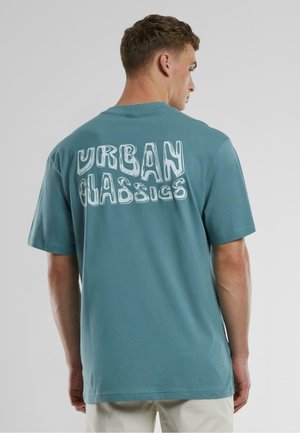 Teal puuvillane T-särk, millel on ümar kaelus, lühikesed varrukad ja julge valge "URBAN CLASSICS" grafika seljal.