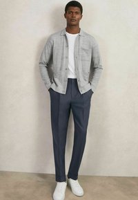 Grau cardigan mit Knopfverschluss und zwei vorderen Taschen, darunter ein weißes T-Shirt, kombiniert mit navyblauen Anzughosen und weißen Sneakers.
