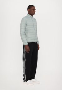 Light gray puffer jacket with a high collar, vertical quilted pattern, and a front zipper. Paired with black sweatpants featuring white stripes.