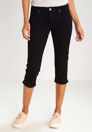 Mujer con pantalones negros ajustados y cortos y zapatillas casuales rosa claro sobre un fondo liso.