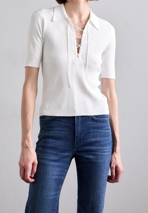 Kvinde iført en hvid ribstrikket polo med snørelukning i halsen og korte ærmer, kombineret med mørkeblå jeans foran en ensfarvet baggrund.