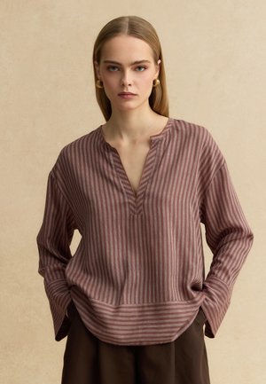 Jonge vrouw met stijl blond haar, goudkleurige oorringen en een losse bruin gestreepte blouse met lange mouwen, met haar handen in de zakken.