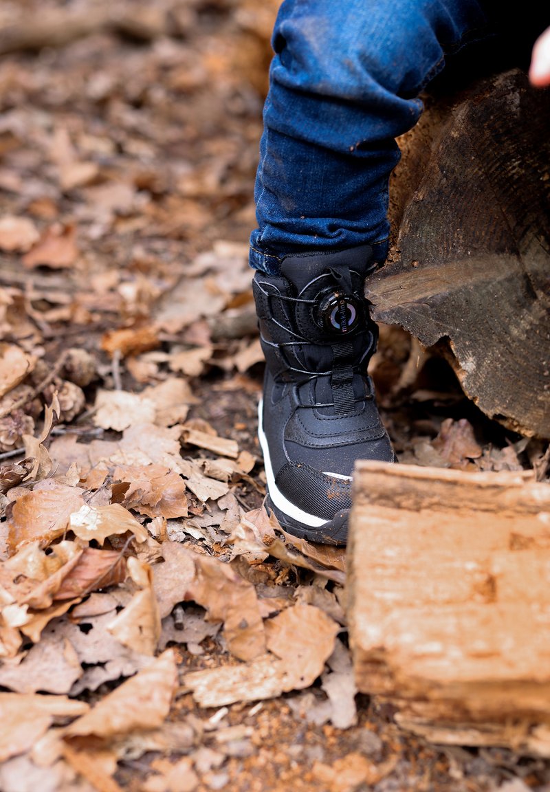 Čierna zimná topánka má textúrovaný zvršok, kruhový systém šnurovania a bielu podrážku. Nachádza sa na zemi pokrytej suchými listami a drevom.