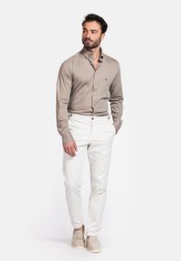Lange mouwen beige shirt met een button-down kraag en een borstzak, gecombineerd met witte broek met gouden knoopaccenten. Beige schoenen maken de look compleet.