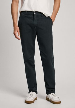 Man draagt donkere casual broek, witte sneakers met bruine zolen en een wit t-shirt, staand met één hand in de zak.