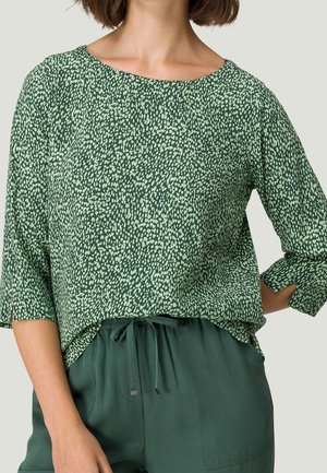 Kobieta w zielonej bluzce w wzory z rękawami o długości do łokcia oraz zielonych spodniach z wiązaniem, stojąca z jedną ręką na biodrze.