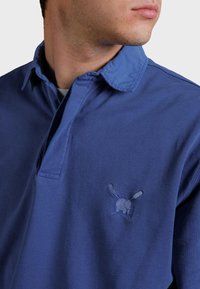 Kungsblå polo skjorta med knäppkrage, försedd med en broderad logotyp på bröstet och ett mjukt, texturerat tyg.